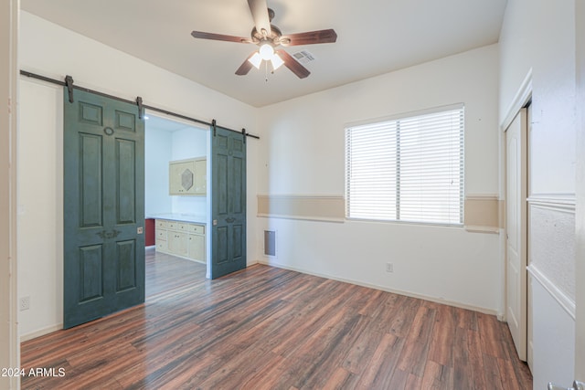 unfurnished bedroom featuring a barn door, ceiling fan, ensuite bathroom, and dark hardwood / wood-style flooring