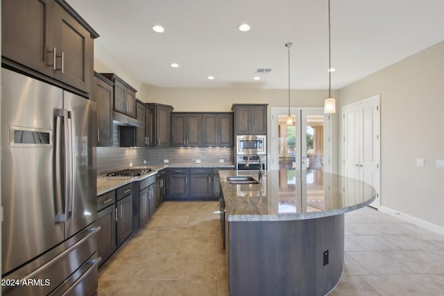 kitchen with dark brown cabinetry, hanging light fixtures, stainless steel appliances, light stone counters, and a center island with sink