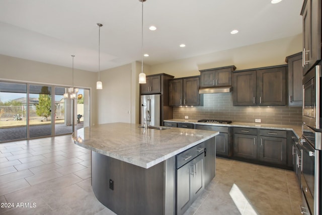 kitchen with a kitchen island with sink, hanging light fixtures, appliances with stainless steel finishes, dark brown cabinets, and light stone counters