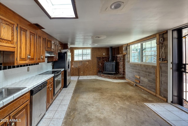 kitchen featuring brown cabinetry, a wood stove, stainless steel appliances, tile counters, and under cabinet range hood