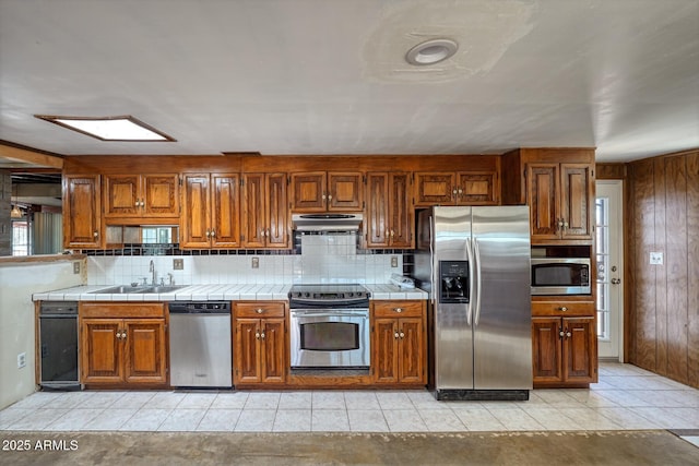 kitchen featuring a sink, under cabinet range hood, stainless steel appliances, brown cabinetry, and tile counters