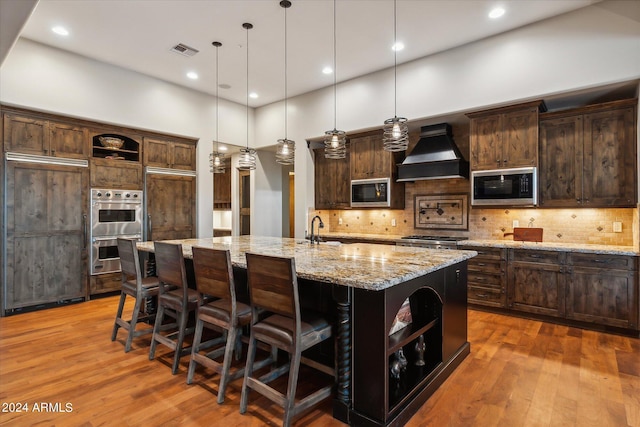 kitchen with built in microwave, a spacious island, hanging light fixtures, custom range hood, and light stone countertops