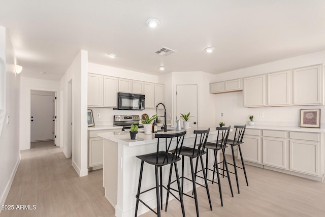 kitchen featuring light countertops, white cabinetry, an island with sink, black microwave, and stainless steel electric range