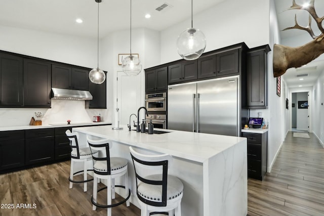 kitchen featuring dark wood-type flooring, sink, decorative light fixtures, a center island with sink, and stainless steel appliances