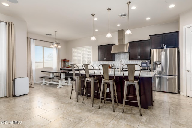 kitchen featuring visible vents, a kitchen island with sink, stainless steel refrigerator with ice dispenser, backsplash, and wall chimney range hood