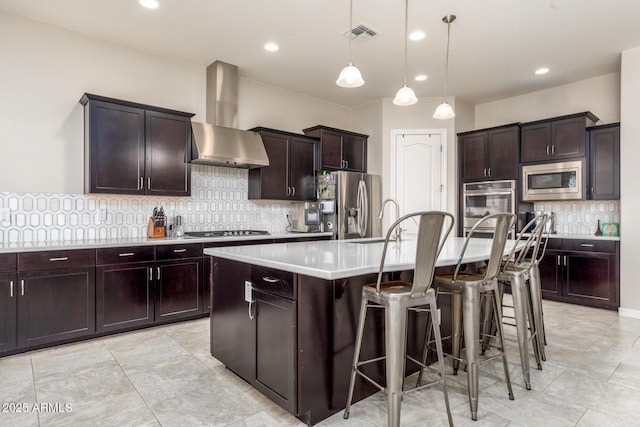 kitchen with an island with sink, stainless steel appliances, light countertops, a kitchen bar, and wall chimney range hood