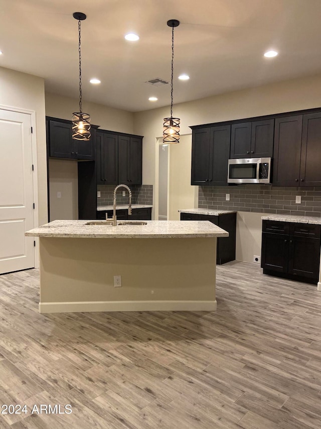 kitchen featuring decorative backsplash, a kitchen island with sink, hanging light fixtures, sink, and light wood-type flooring