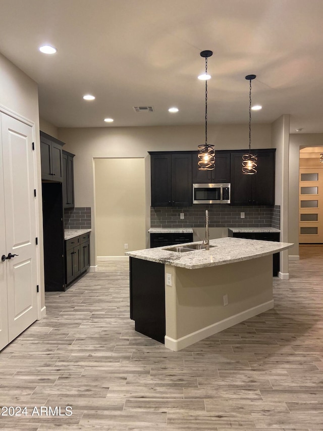 kitchen with sink, an island with sink, backsplash, hanging light fixtures, and light stone counters