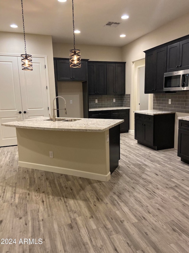 kitchen with decorative backsplash, an island with sink, light hardwood / wood-style flooring, sink, and decorative light fixtures