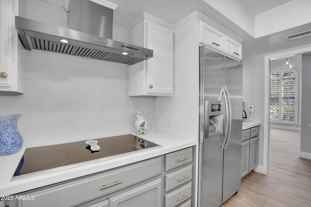 kitchen featuring stainless steel fridge with ice dispenser, black electric stovetop, wall chimney range hood, light wood-style flooring, and visible vents
