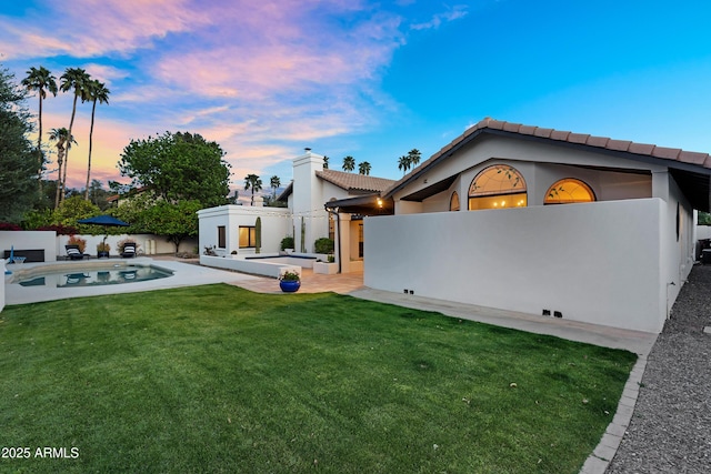 back of house at dusk featuring a yard, a tile roof, fence, stucco siding, and a fenced in pool