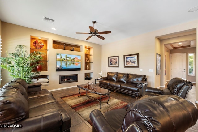 living area with ceiling fan, a glass covered fireplace, built in shelves, baseboards, and visible vents