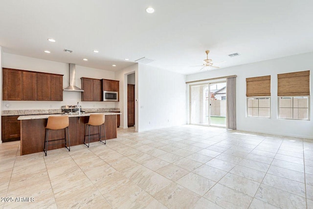 kitchen featuring wall chimney range hood, a breakfast bar, stainless steel microwave, light countertops, and an island with sink