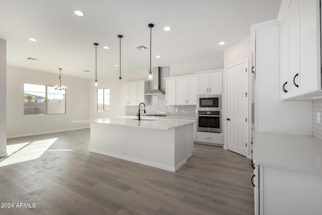 kitchen featuring a kitchen island with sink, white cabinets, wall chimney range hood, light wood-type flooring, and appliances with stainless steel finishes