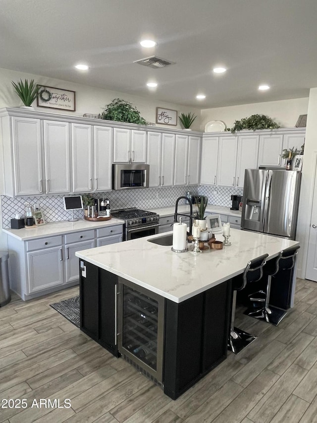 kitchen with sink, appliances with stainless steel finishes, a kitchen island with sink, wine cooler, and light stone countertops