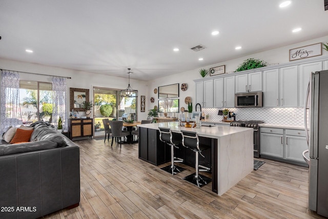 kitchen with appliances with stainless steel finishes, an island with sink, decorative backsplash, hanging light fixtures, and light hardwood / wood-style floors