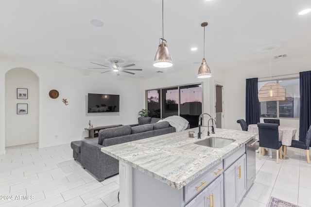 kitchen featuring light stone counters, ceiling fan, a kitchen island with sink, sink, and pendant lighting