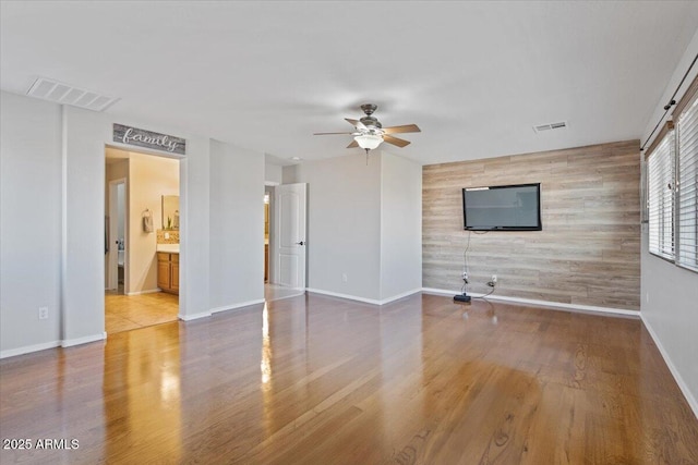 unfurnished living room featuring visible vents, a ceiling fan, wood finished floors, wooden walls, and an accent wall