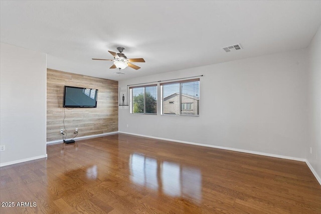 unfurnished living room featuring visible vents, an accent wall, ceiling fan, baseboards, and wood finished floors