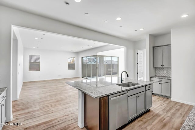 kitchen with an island with sink, sink, stainless steel dishwasher, light stone countertops, and light wood-type flooring