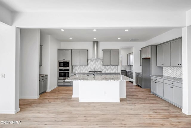 kitchen with built in microwave, wall chimney exhaust hood, stainless steel oven, a center island with sink, and light stone countertops