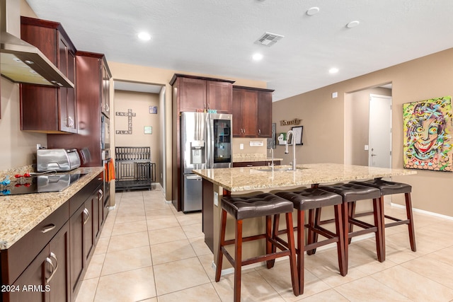 kitchen with stainless steel refrigerator with ice dispenser, sink, wall chimney exhaust hood, and an island with sink