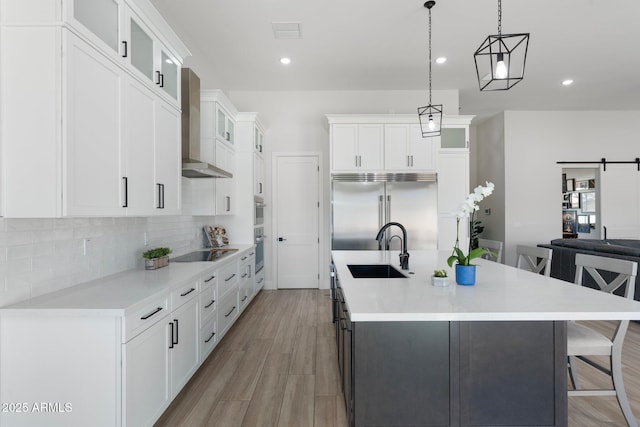 kitchen with built in refrigerator, decorative light fixtures, wall chimney range hood, sink, and a barn door