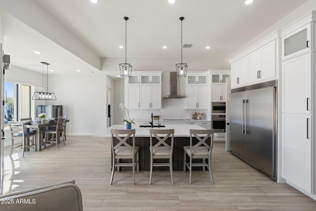 kitchen with appliances with stainless steel finishes, wall chimney range hood, decorative light fixtures, white cabinets, and a center island with sink