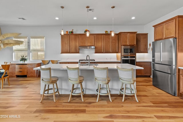 kitchen featuring appliances with stainless steel finishes, light wood-type flooring, a kitchen island with sink, sink, and hanging light fixtures