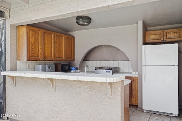 kitchen with freestanding refrigerator, black microwave, tile countertops, and brown cabinets