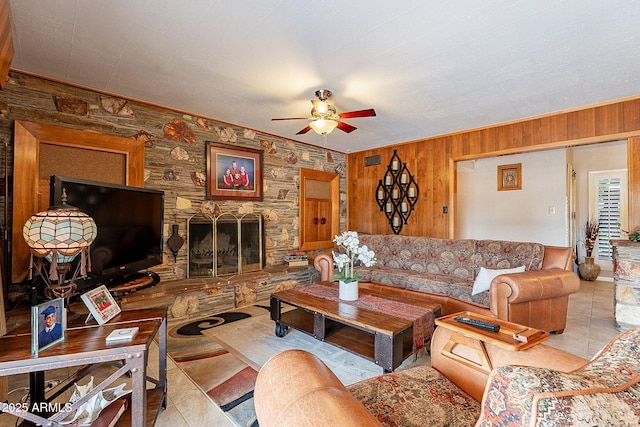 tiled living room featuring ceiling fan, wood walls, and a stone fireplace