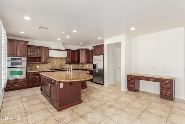 kitchen featuring light stone countertops, premium range hood, tasteful backsplash, an island with sink, and stainless steel appliances