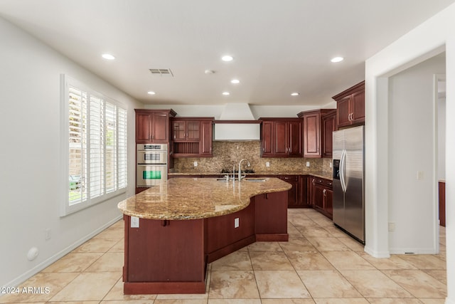 kitchen featuring backsplash, stainless steel appliances, premium range hood, an island with sink, and light stone countertops