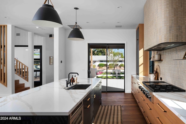 kitchen with wall chimney range hood, hanging light fixtures, an island with sink, dark wood-type flooring, and sink