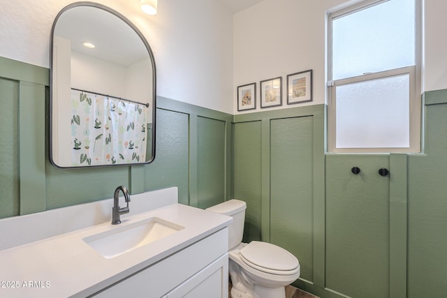 bathroom featuring curtained shower, a decorative wall, toilet, wainscoting, and vanity