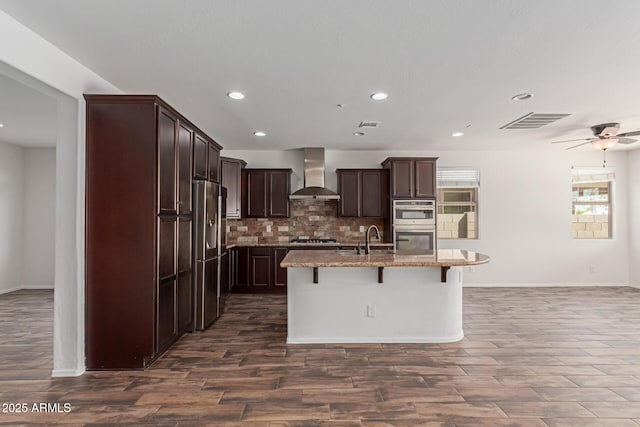 kitchen with light stone counters, visible vents, stainless steel appliances, a kitchen bar, and wall chimney exhaust hood