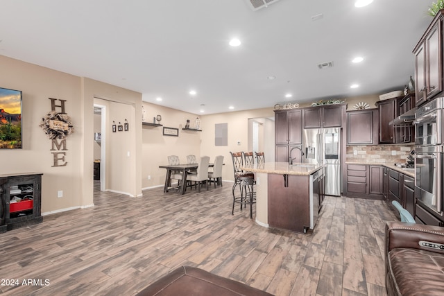 kitchen with a kitchen breakfast bar, dark brown cabinets, a kitchen island with sink, hardwood / wood-style flooring, and light stone counters