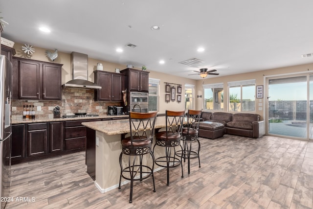 kitchen featuring a center island, a breakfast bar, ceiling fan, wall chimney range hood, and light wood-type flooring