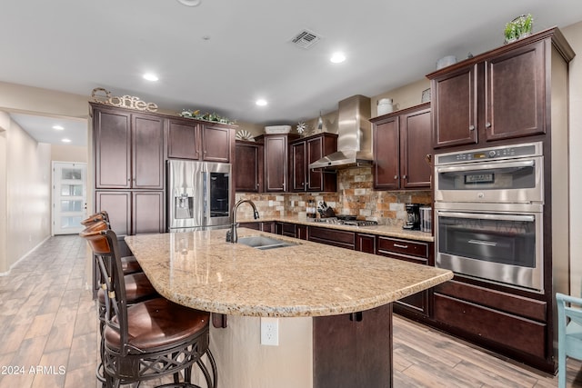 kitchen featuring an island with sink, appliances with stainless steel finishes, wall chimney range hood, and a kitchen bar