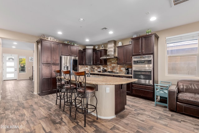 kitchen with stainless steel appliances, light wood-type flooring, wall chimney exhaust hood, and a kitchen breakfast bar