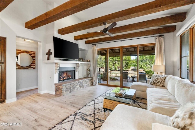 living room featuring baseboards, wood finished floors, ceiling fan, a stone fireplace, and beam ceiling