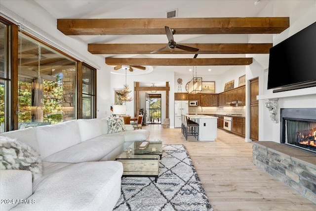 living area featuring beamed ceiling, a warm lit fireplace, ceiling fan, light wood-type flooring, and visible vents