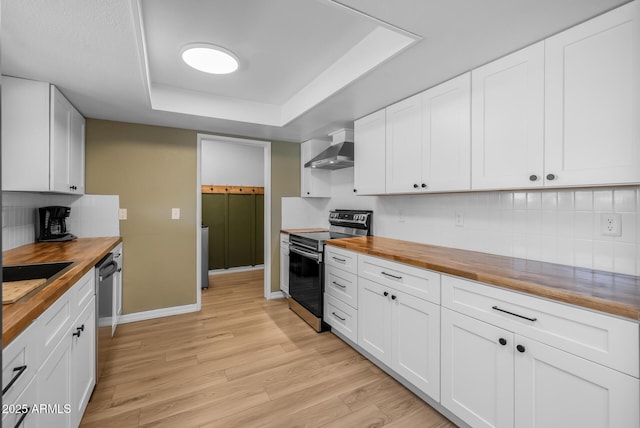 kitchen featuring stainless steel appliances, wall chimney exhaust hood, wood counters, a tray ceiling, and light wood-style flooring