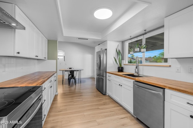 kitchen featuring wall chimney range hood, a sink, butcher block countertops, stainless steel appliances, and a tray ceiling