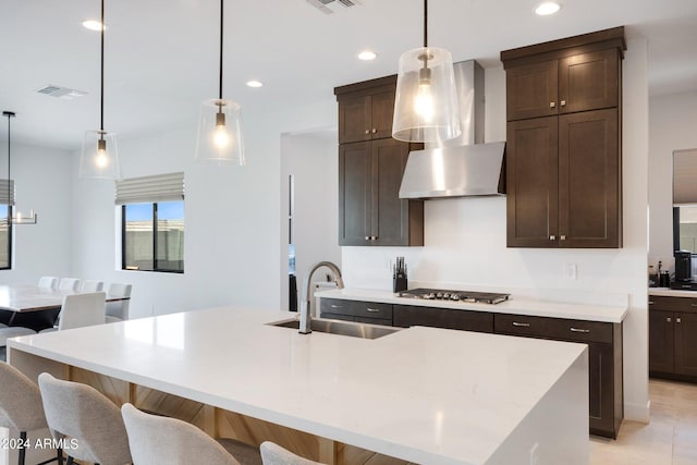 kitchen featuring wall chimney range hood, sink, a center island with sink, and hanging light fixtures