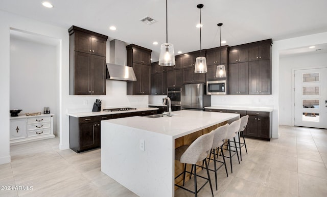 kitchen featuring hanging light fixtures, appliances with stainless steel finishes, a kitchen island with sink, wall chimney exhaust hood, and sink