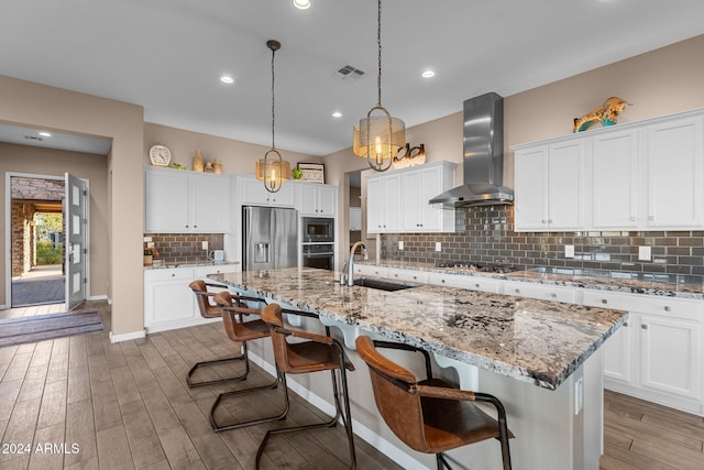 kitchen with stainless steel appliances, hardwood / wood-style flooring, wall chimney exhaust hood, and a kitchen island with sink