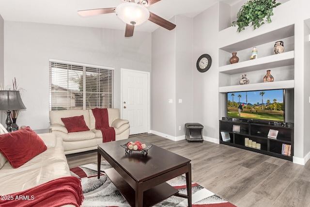 living room with built in shelves, a towering ceiling, a ceiling fan, wood finished floors, and baseboards