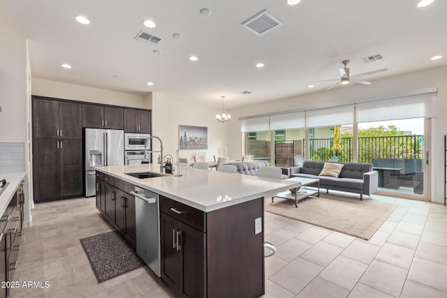 kitchen with ceiling fan with notable chandelier, sink, an island with sink, appliances with stainless steel finishes, and dark brown cabinets