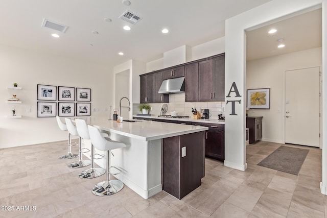 kitchen featuring sink, decorative backsplash, an island with sink, dark brown cabinets, and stainless steel gas cooktop